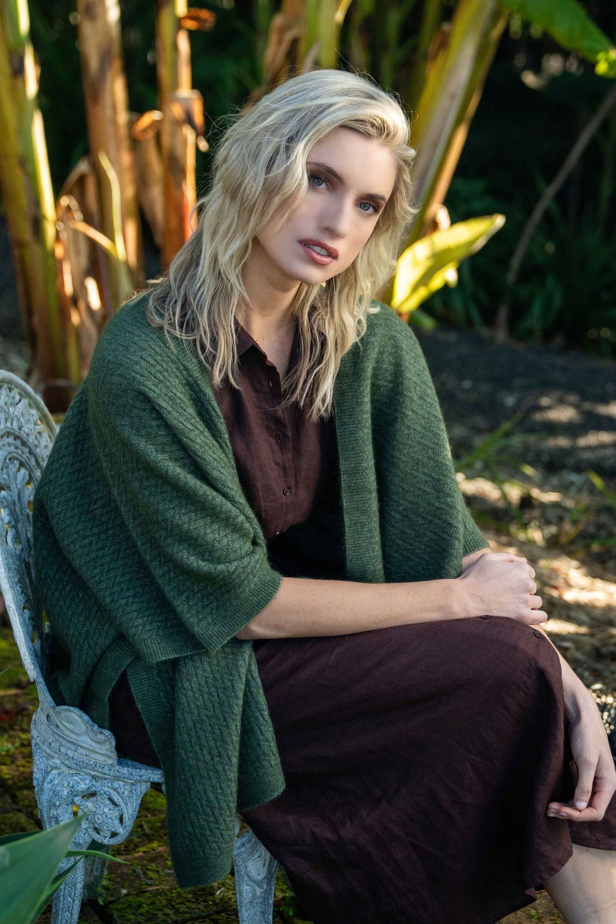 A woman sits on a white garden chair outdoors, wearing the Zig Zag Textured Possum Merino Cape in Moss by Koru over a dark brown dress. The background features lush green foliage and warm sunlight.