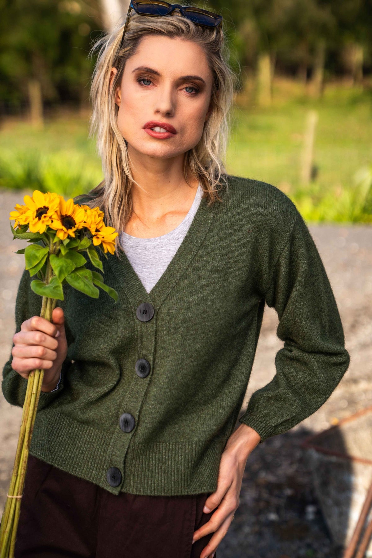 A woman outdoors wearing the V Neck Possum Merino Cardigan in Moss by Koru over a light gray top, holding a bouquet of yellow sunflowers. The background shows a blurred natural setting with greenery.