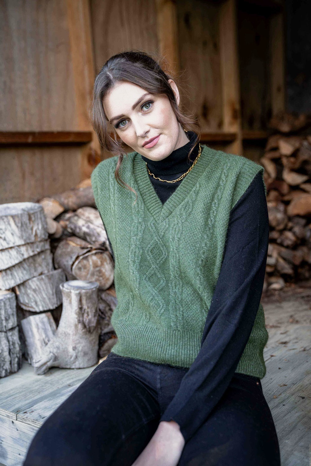 Woman wearing the Cable Possum Merino Vest in Moss by Koru sitting in front of stacked firewood.