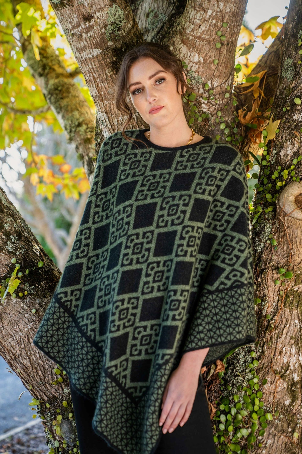 Woman wearing the Diamond Possum Merino Poncho in Black/Moss by Koru standing against a tree with autumn leaves.