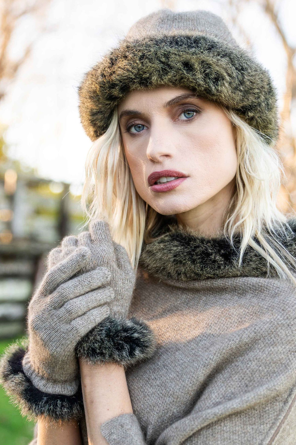 A woman the Fur Trim Possum Merino Gloves in Mocha/Snowtip by Koru with matching Beanie and Poncho, photographed outdoors with a softly blurred background.