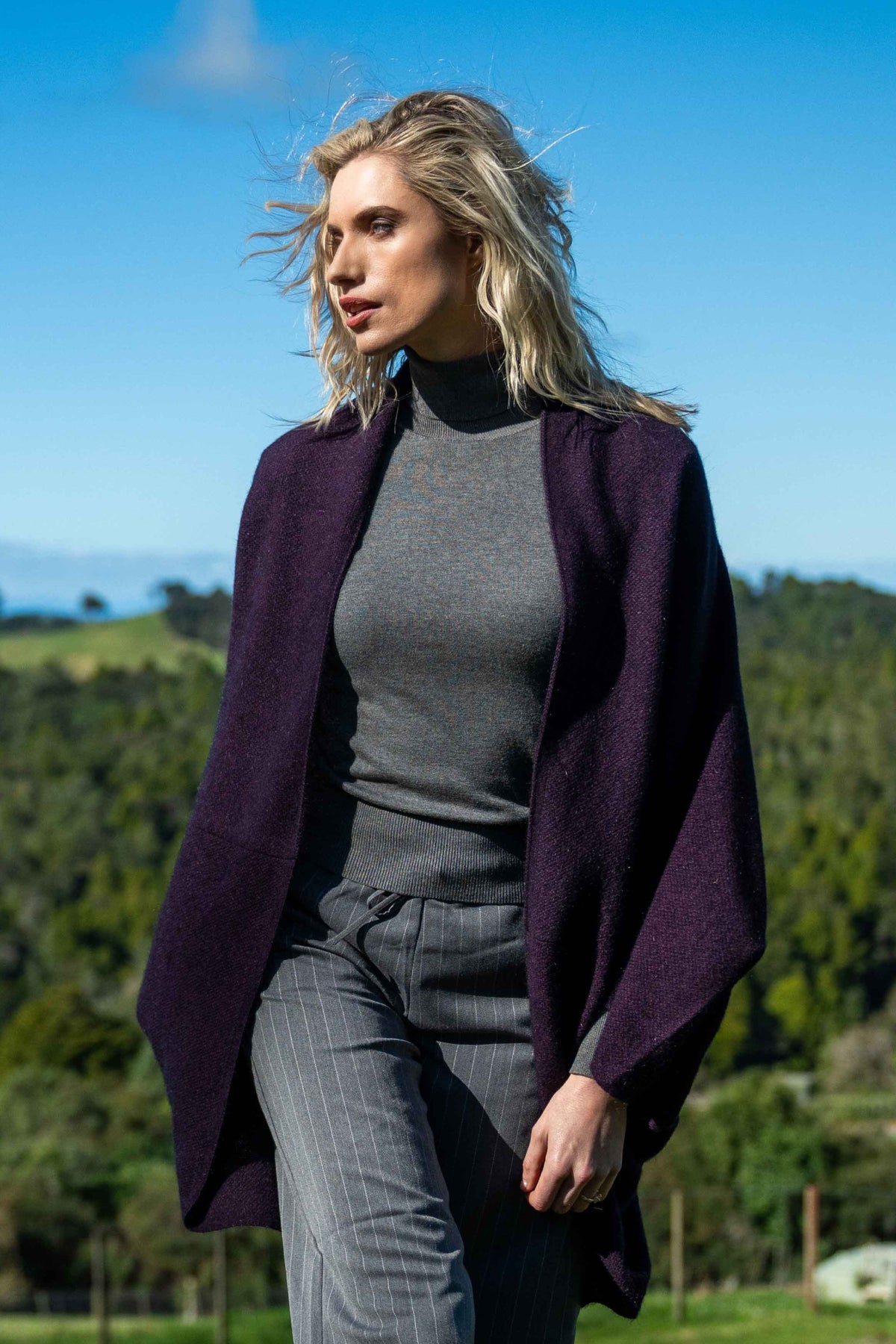 A woman outdoors wearing the Moss Stitch Possum Merino Shrug in Grape by Koru over a gray mock-neck top and gray pinstripe trousers, set against a background of green hills and blue sky.