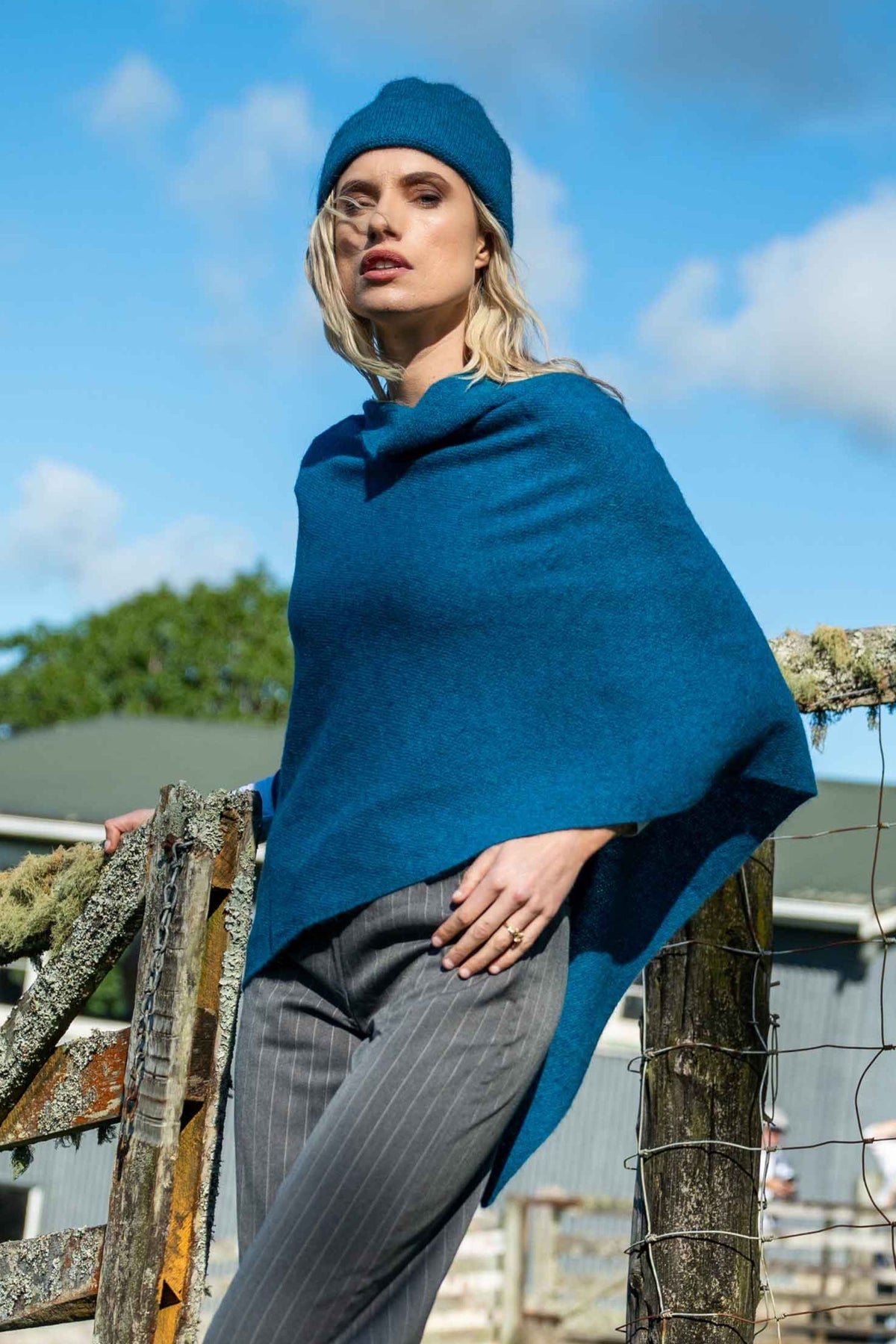 A woman wearing the Two Way Possum Merino Poncho in Pacific by Koru, matching beanie, and gray pinstripe trousers, posing outdoors against a rustic wooden fence under a blue sky.