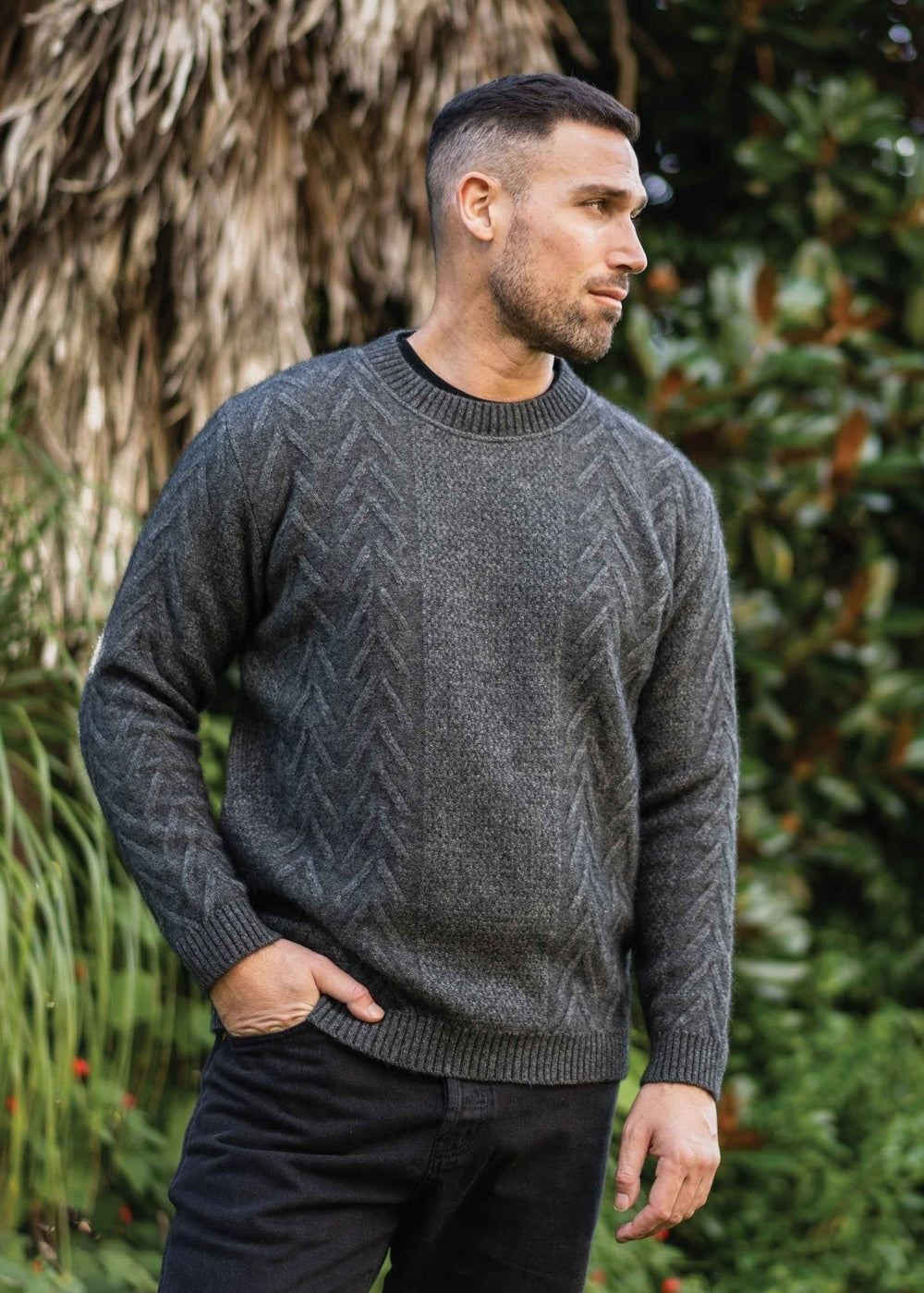 Man wearing the Possum Merino Aran Crew Neck in Grey by Koru standing outdoors with greenery in the background