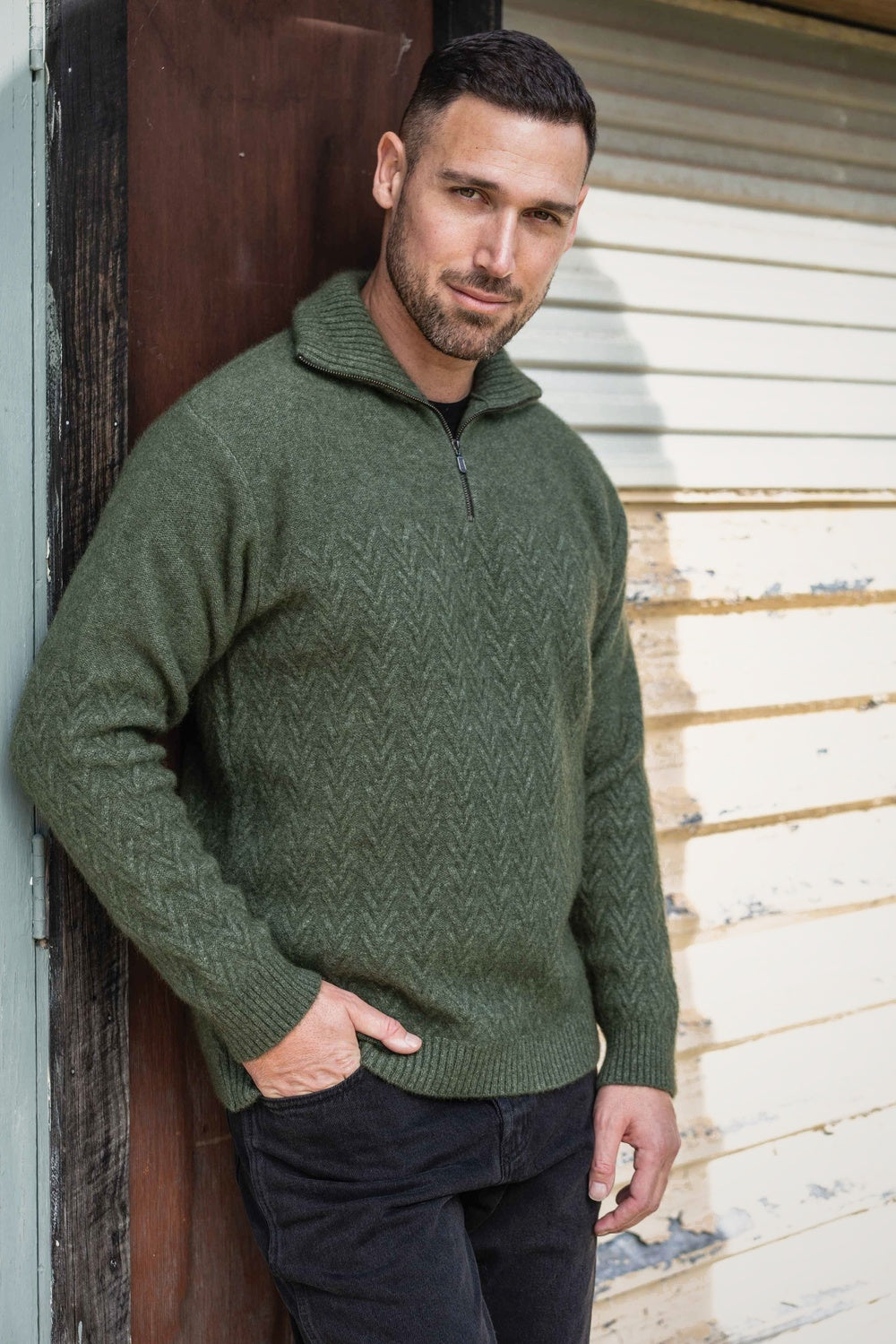 Man wearing the Chevron Zip Possum Merino Jumper in Moss by Koru leaning against a wooden door.
