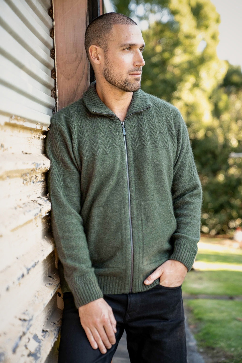 Man wearing the Chevron Zip Possum Merino Jacket in Moss by Koru leaning against a wall outdoors.