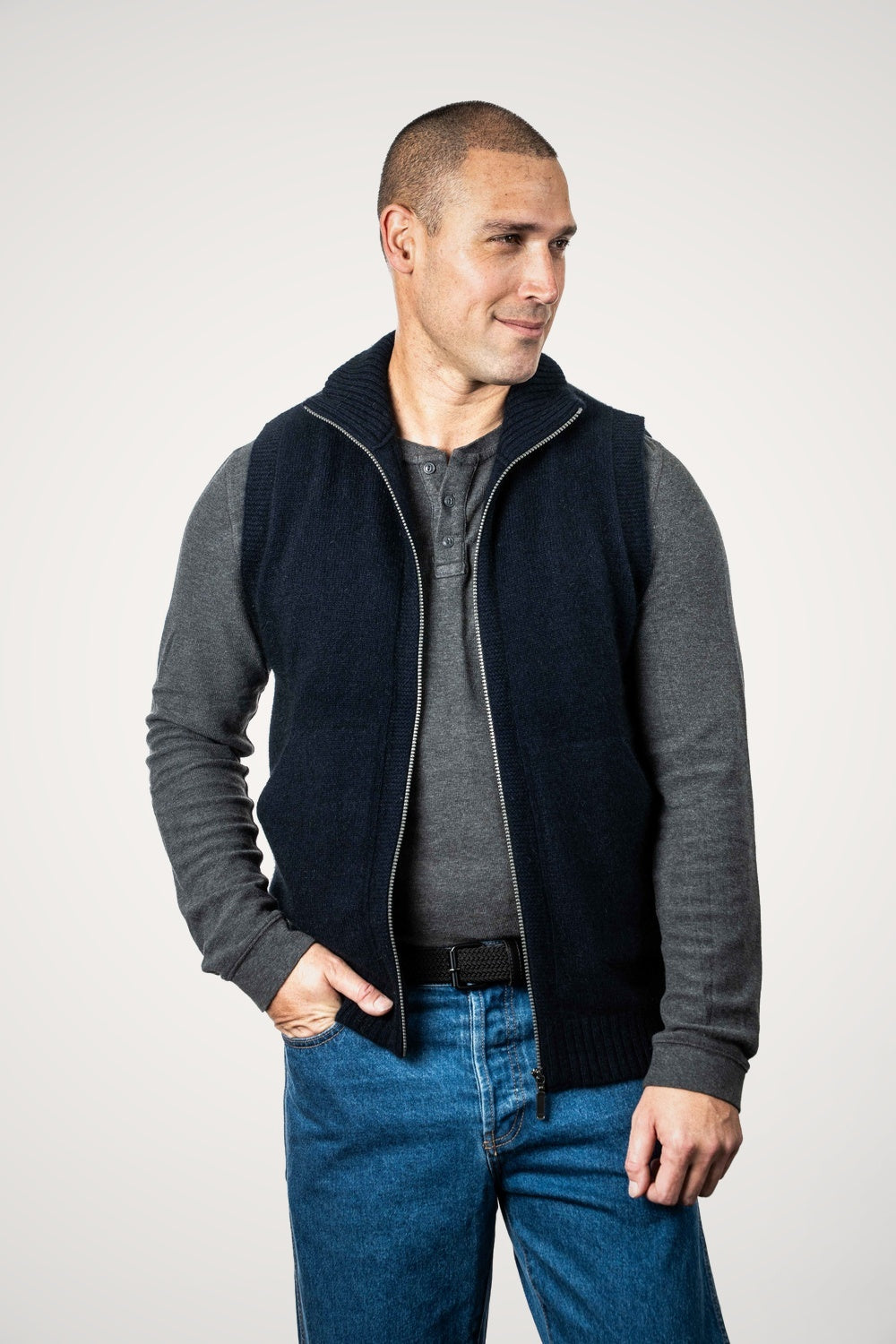 Man wearing the Zip Vest with Collar Possum Merino in Navy by Koru over a gray long-sleeve shirt and blue jeans on a white background