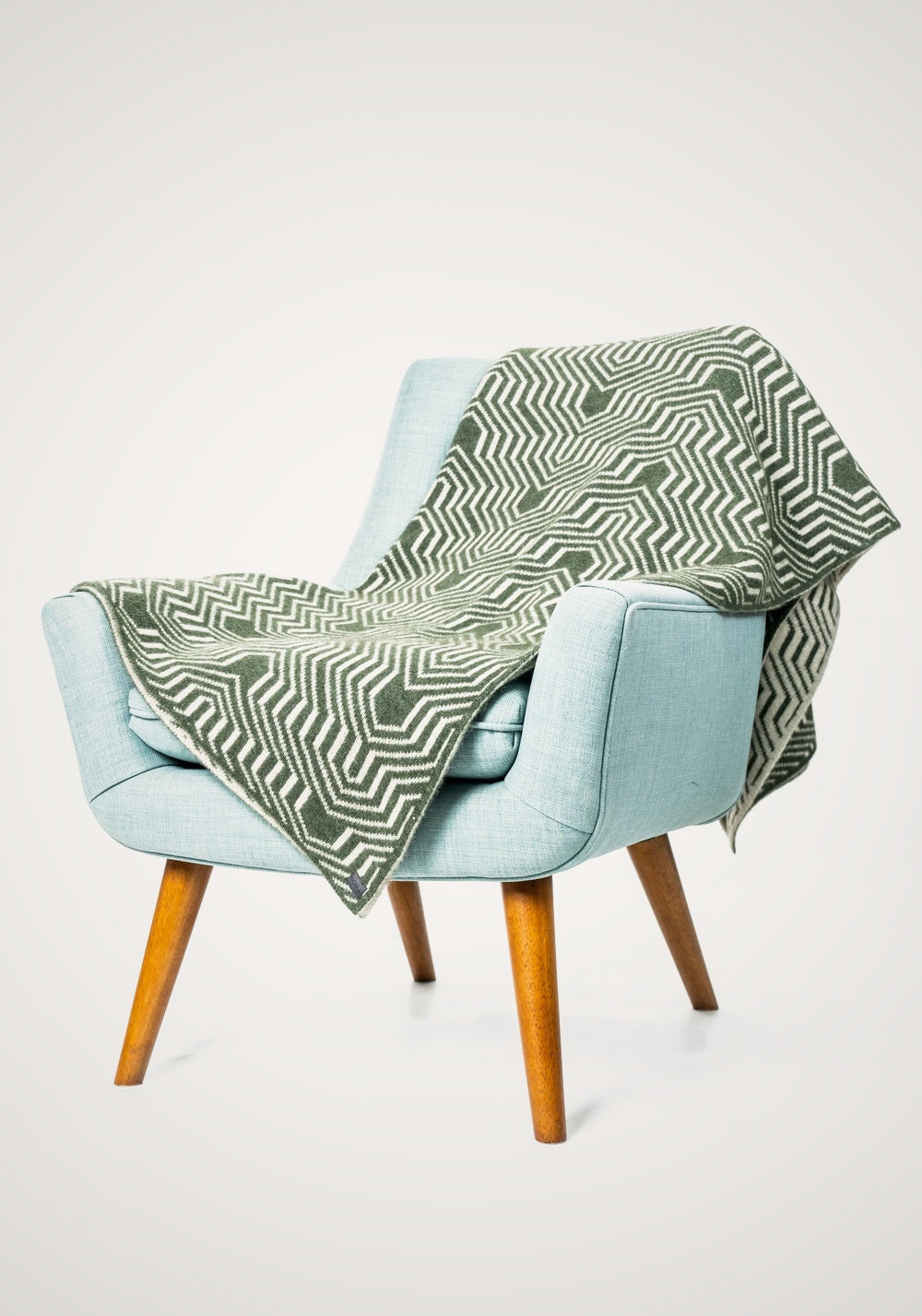 Light blue chair with the Patterned Possum Merino Throw in Moss/White by Koru draped over iit on a white background