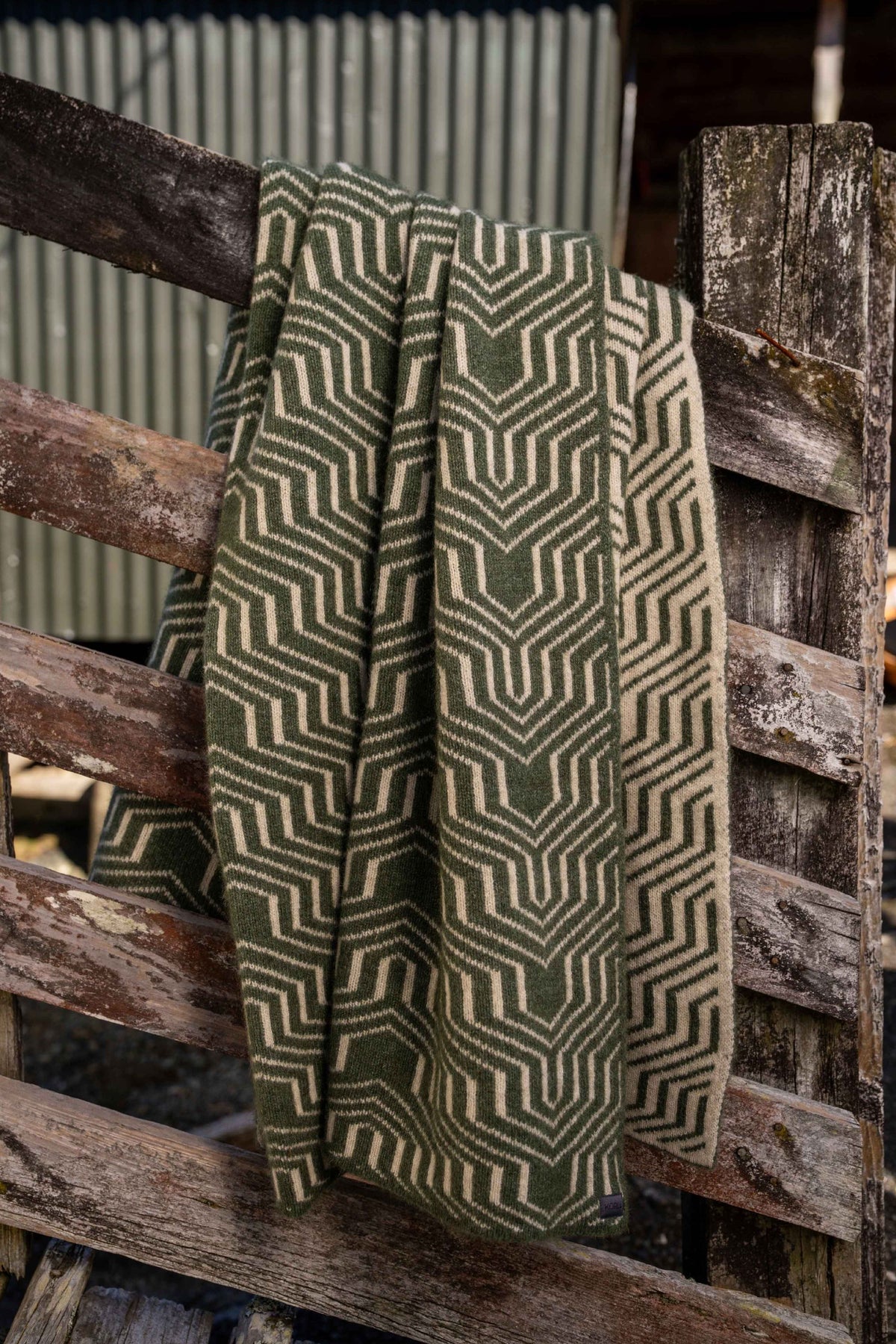 The Patterned Possum Merino Throw in Moss/White by Koru draped over rustic, wooden fence rails