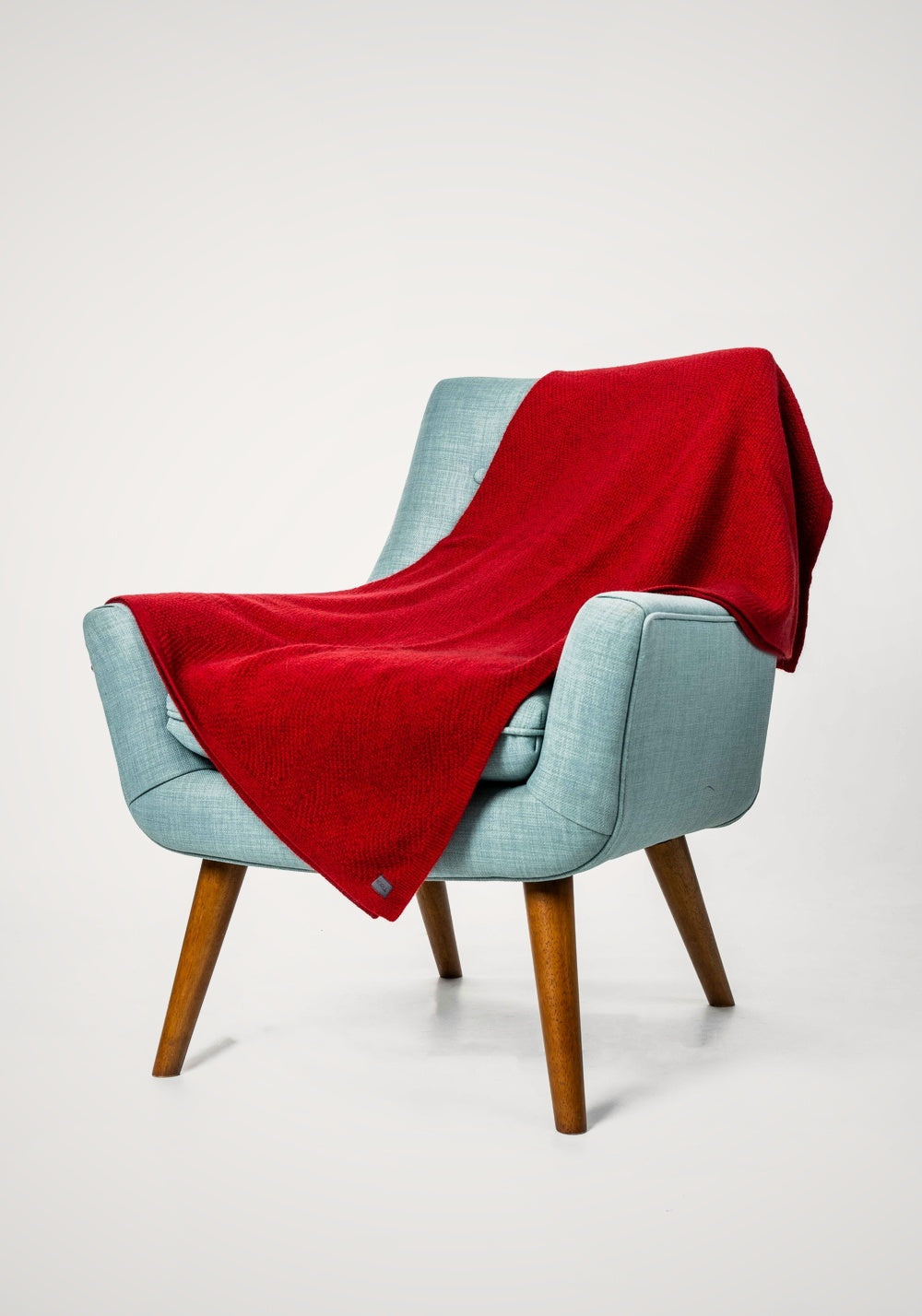 Light blue chair with the Plated Herringbone Possum Merino Throw in Rata/Red by Koru draped over it on a white background