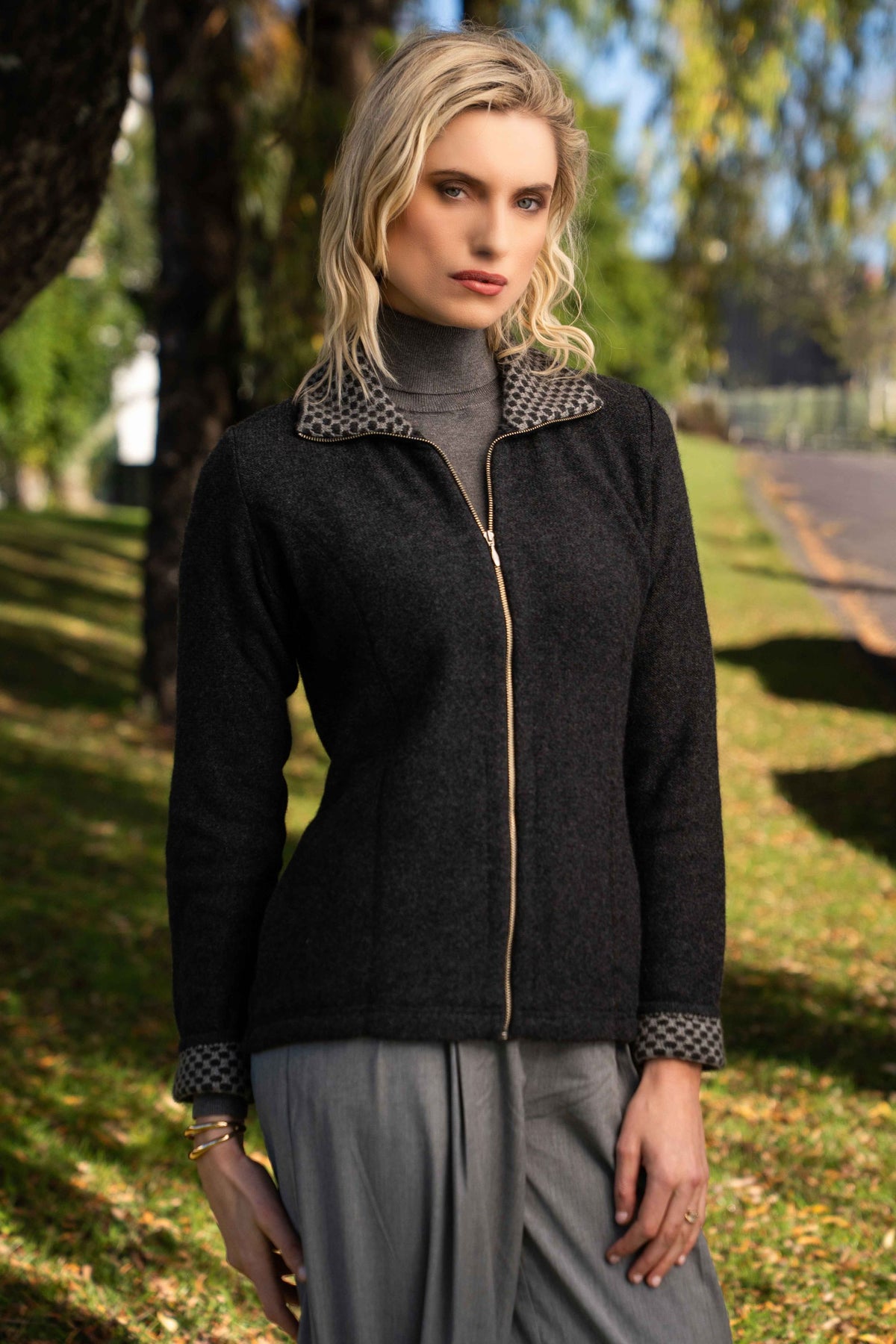 A woman standing outdoors in a park wearing the Contrast Trim Possum Merino Jacket in Black Marl/Mist by Trilogy, paired with gray trousers.