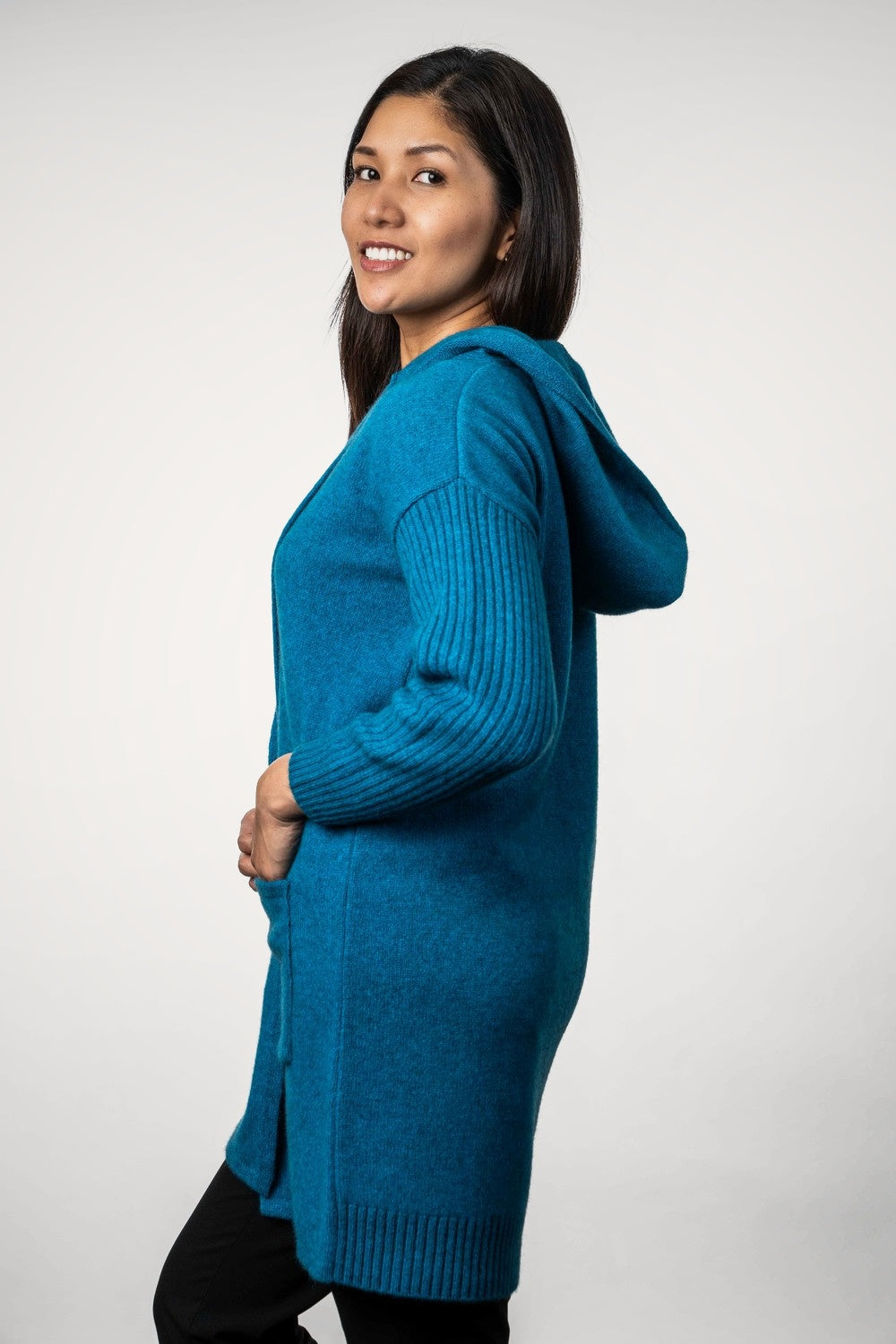 Woman wearing the Hooded Longline Possum Merino Cardigan in Teal by Trilogy against a white background - side view