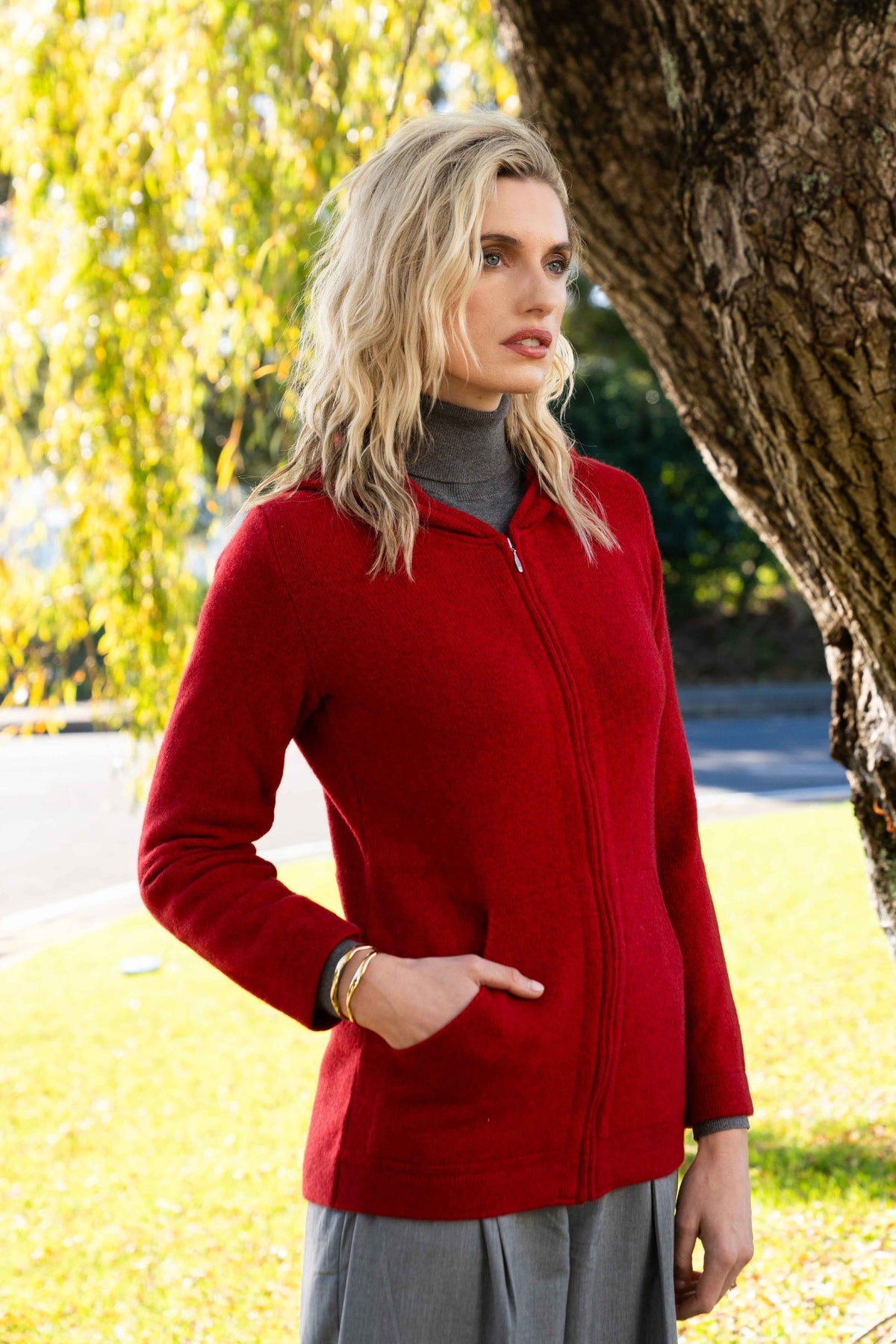 A woman wearing the the Hooded Zip Possum Merino Jacket in Scarlet by Trilogy over a gray mock neck top and gray skirt, standing outdoors next to a large tree trunk with green grass and foliage in the background.